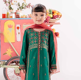 Child in a green embroidered outfit standing in front of a colorful cart with fruits.