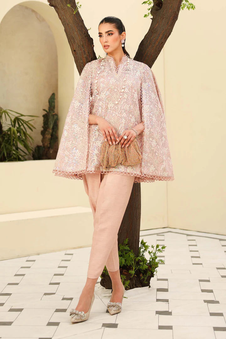 Woman in a light pink traditional outfit standing outdoors with plants and a tree in the background.