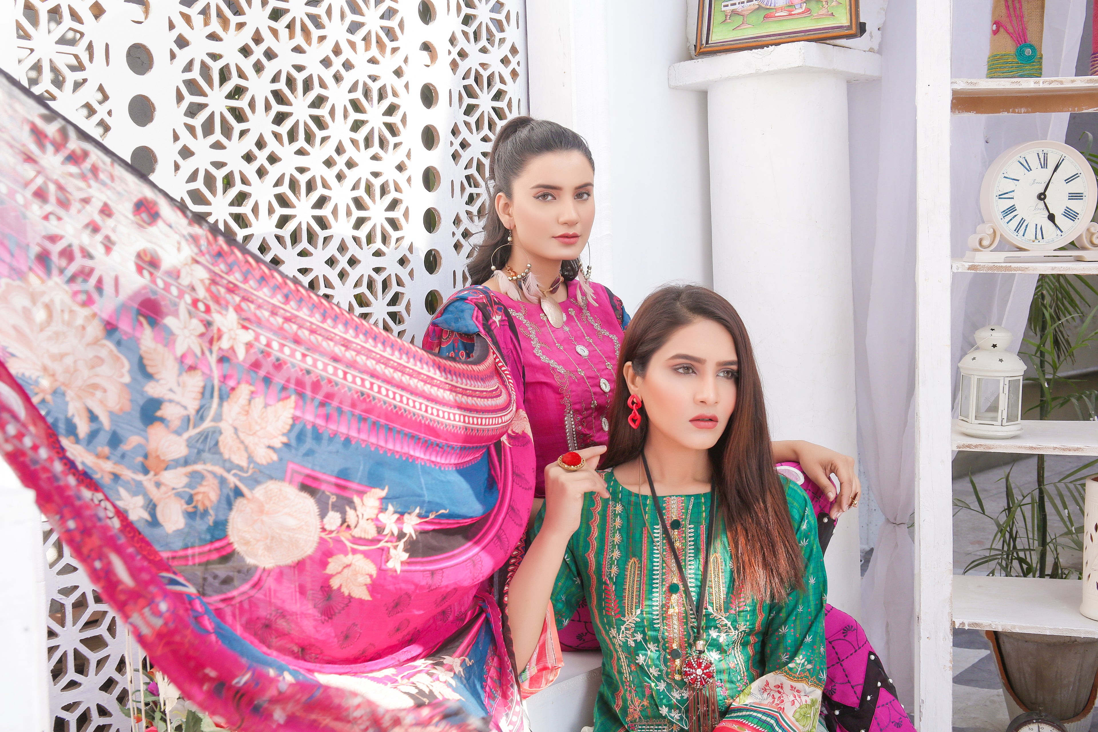 Two women in colorful traditional outfits with a decorative wall and shelf in the background.